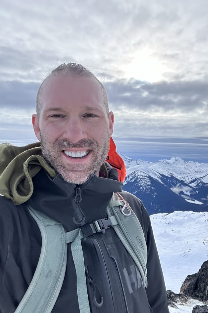 Dave standing on top of a mountain in front of a mountain range and sun through the clouds.