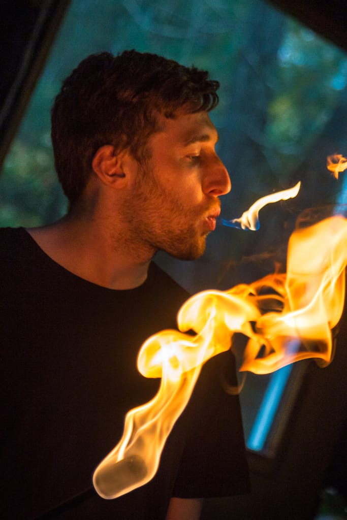 Taylor in dark setting with stream of fire coming from his lips, lit torch in foreground.